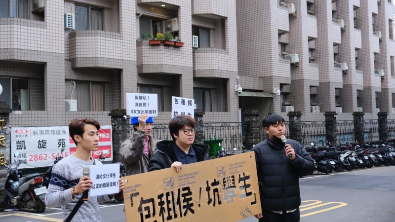 中国文化大学学生在出租套房凯旋苑前高举标语抗议，呼吁侯友宜正面回应凯旋苑涨租议题，让房租合理化。