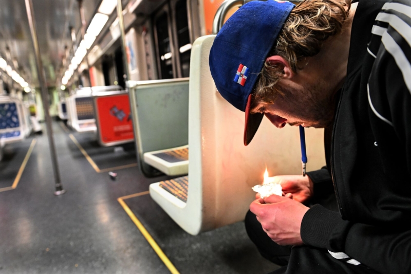 Seated in a subway train, a young man with dark blond hair and a baseball cap smokes fentanyl.