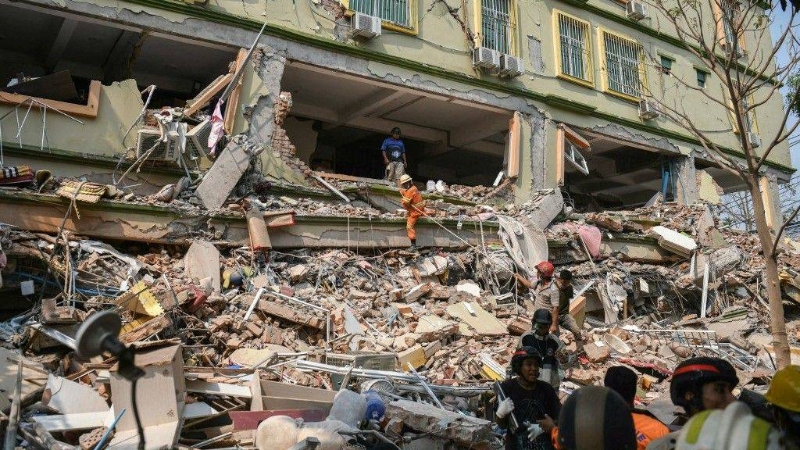 在曼德勒郊区一所倒塌的私立中学废墟前，搜救人员正努力搜寻被困者。 Thai PBS World